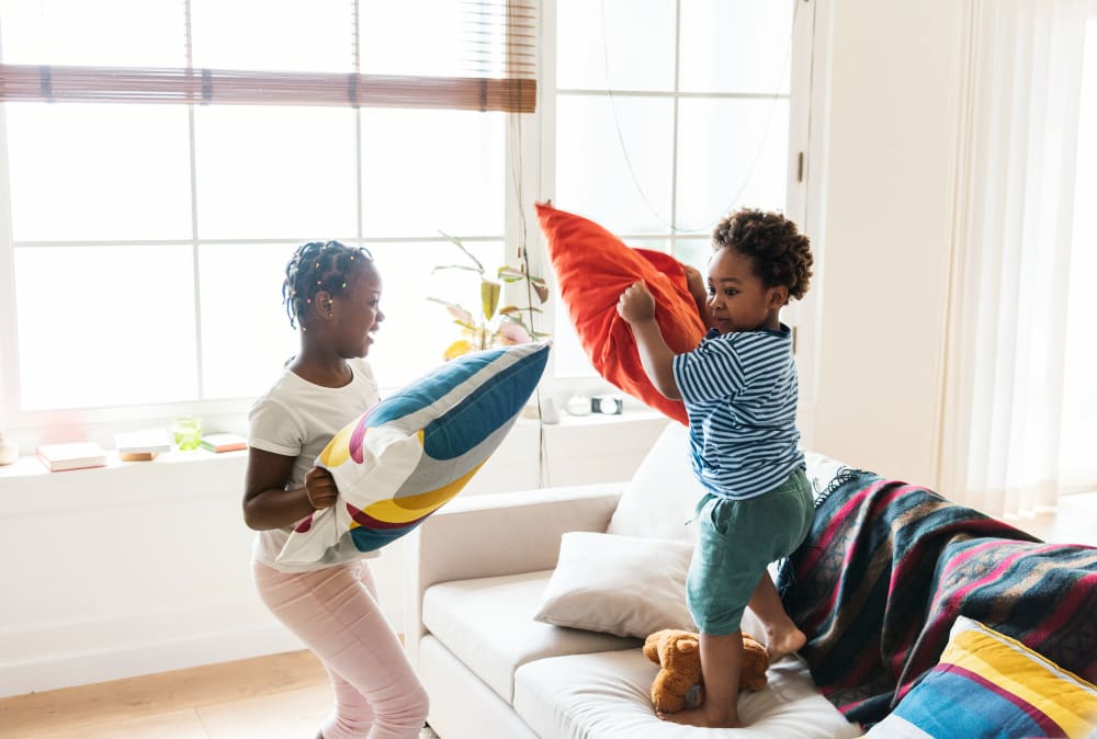 Two children mid pillow-fight on a sofa in a bright living room, both laughing.