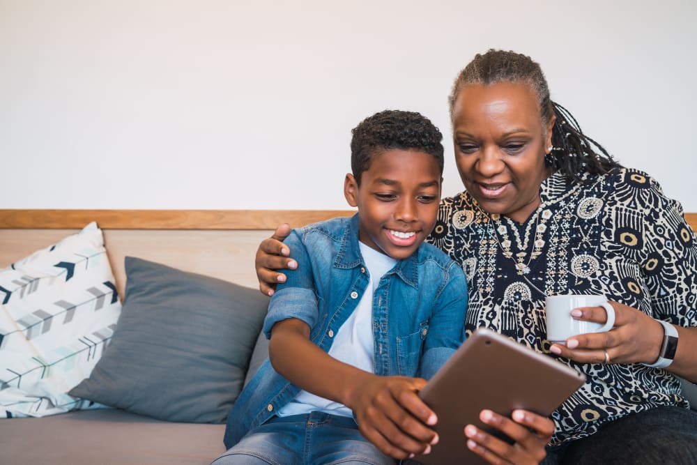 A grandmother with her arm around her grandson, both smiling at a tablet screen held between them.