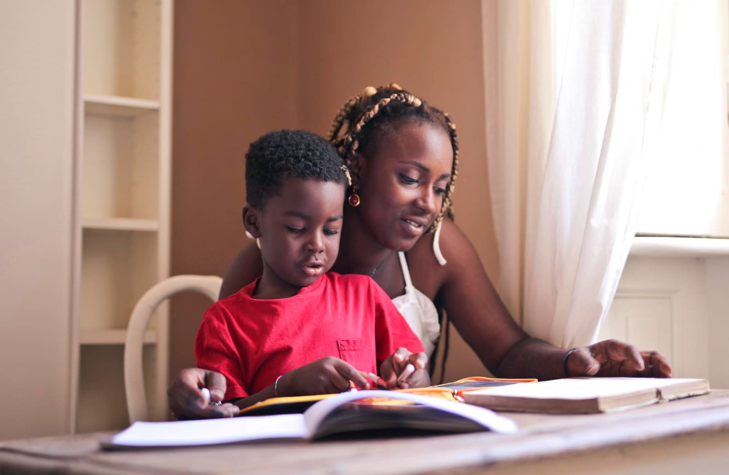 A mother and her young son reading together at a table in warm daylight, his hand on the page.