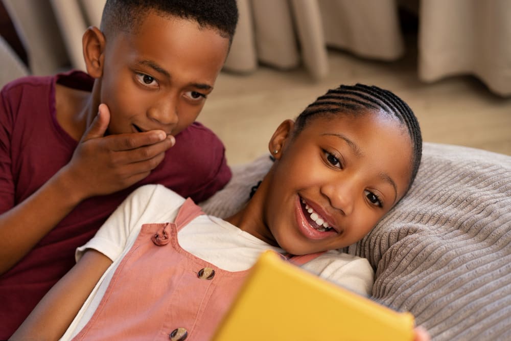 A brother and sister lying back together, sharing something on a tablet and grinning.